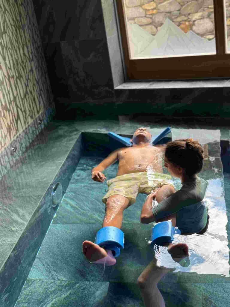 Séance d’ostéopathie aquatique avec Laetitia Landais-Maillard, un homme flotte avec des flotteurs pendant que l’ostéopathe mobilise sa jambe dans l’eau.